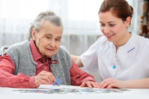 Ältere Frau sitzt mit einer Pflegeperson an einem Tisch und spielt ein Puzzle.